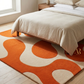 Orange and beige patterned rug on a wooden floor with a bed and lamp in the background.
