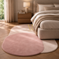 A modern oval area rug in dusty pink and cream, styled in a warm, sunlit bedroom next to a beige bed and wooden nightstand.