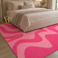 A pink and light pink abstract wavy patterned area rug on a light wood floor in a modern bedroom setting with a beige bed.