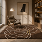 A large brown area rug with a white abstract line pattern and a small yellow accent, styled in a modern, sunlit reading room with a mid-century armchair, floor lamp, and bookshelves.