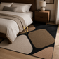 A modern area rug with an abstract organic pattern in black, beige, and brown, styled on a light wood floor in a bedroom next to a bed and a wooden nightstand.