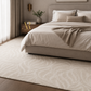 A large beige area rug with a subtle wavy pattern covers most of the floor in a modern, neutral-toned bedroom. The room features a bed with beige bedding, a wooden nightstand, and soft natural light coming from a window on the left.