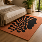 An orange area rug with a large black abstract leaf and face profile design, styled in a modern bedroom next to a bed and a potted plant.