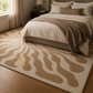 A large area rug with a beige and cream wavy, organic pattern is shown on a hardwood floor in a styled bedroom setting with a bed, pillows, and a nightstand.