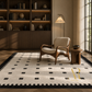 A large rectangular area rug with a cream base, black geometric grid pattern, and black scalloped border, styled in a warm, modern living room with a wooden armchair, side table, and bookshelves.