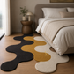 A modern wavy-shaped area rug in black, mustard yellow, and cream, styled on a light wood floor in a bedroom next to a bed.