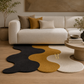 A modern wavy area rug in black, mustard yellow, and cream, styled on a light wood floor in front of a cream-colored sofa with decorative pillows.