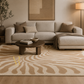 A modern living room scene featuring a large beige and tan patterned area rug under a light beige sectional sofa with brown accent pillows and a matching throw blanket.