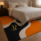 A modern area rug with an abstract wavy pattern in black, orange, cream, and gray, styled on a wooden floor in front of a bed with beige linens. The rug features a gold 'AR' monogram and 'Artisti Rugs' branding.