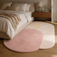 Two overlapping oval area rugs on a wooden floor in a styled bedroom. The front rug is dusty pink, and the rear rug is cream with a gold monogram logo.