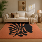 A large rectangular area rug in terracotta orange with a bold black abstract botanical and profile silhouette design, styled in a modern living room setting in front of a cream sofa.