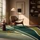 A modern living room scene featuring a large area rug with abstract wavy lines in shades of green, teal, and cream. The room is styled with a mid-century modern wooden armchair, a floor lamp, a small side table with a vase of branches, and a wooden bookshelf in the background.