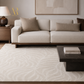 A modern living room scene featuring a large cream and beige patterned area rug, a light beige sofa with matching throw pillows, a dark wood side table with a lamp, and a dark coffee table in the foreground.