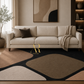 A modern abstract area rug in beige, brown, and black is styled in a living room setting under a cream-colored sofa with decorative pillows and a throw blanket.