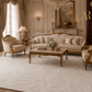 An elegant, luxurious living room featuring a cream tufted sofa, matching armchair, ornate gold-framed coffee table, and a large cream patterned area rug. The room is decorated with gold paneling, a crystal chandelier, and a large mirror.