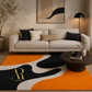 A modern living room scene featuring a large abstract area rug with bold orange, black, and cream shapes. The rug has a gold monogram logo 'AR' and the brand name 'Artista Rugs' printed in the lower left.