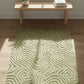 Green patterned rug on a wooden floor with a wooden table and plant in the background.