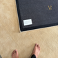 Person standing on a textured beige floor next to a dark rectangular mat with a logo.