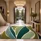 A large rectangular area rug with an abstract leaf-like pattern in shades of green, teal, and cream, displayed on a marble foyer floor. The rug features a gold monogram 'R' in the bottom left corner.