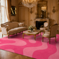 A luxurious pink and magenta wavy patterned area rug in a grand, ornate classical room with gold furniture and a crystal chandelier. A watermark logo for 'Artema Rugi' is visible in the upper left corner.
