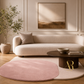 A styled living room scene featuring a cream-colored curved sofa with a matching pillow, a round dusty pink area rug, a dark wood coffee table, and a beige potted plant against a cream paneled wall with a framed abstract painting.