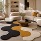 A modern living room featuring a large, abstract wavy area rug in black, cream, and mustard yellow. The room is styled with a curved cream boucle sofa, a round wooden coffee table, and warm natural light streaming in from a window.