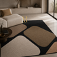A modern abstract area rug with large rounded geometric shapes in beige, taupe, cream, and black, styled in a contemporary living room next to a light beige sofa.