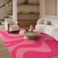 A modern living room scene featuring a prominent pink and light pink wavy patterned area rug. The rug is styled in a contemporary interior with a cream-colored curved sofa, a matching armchair, a round wooden coffee table, and minimalist wall decor.