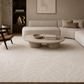 A large beige area rug with a subtle wavy pattern covers the floor in a modern living room setting with a light beige sectional sofa, a round stone coffee table, and a dark wood shelving unit in the background.