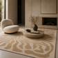 A large area rug with a beige and cream wavy, organic pattern is shown in a modern, minimalist living room setting. The room features a textured cream boucle lounge chair, a round stone coffee table with a vase of dried branches, and a stone fireplace in the background.