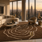A luxury living room scene featuring a large brown shag area rug with a cream concentric circle pattern and a gold monogram logo. The room is styled with a cream curved sofa, a modern accent chair, a marble coffee table, and a gold side table, all set against a backdrop of floor-to-ceiling windows showing a city skyline at sunset.
