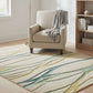 Beige armchair with a gray blanket on a patterned rug in a room with a bookshelf and lamp.