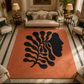 A large rectangular area rug in terracotta orange with a bold black abstract botanical and face profile design, shown in a luxurious, symmetrical living room setting.