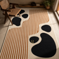A modern area rug with an abstract design featuring black organic shapes and vertical tan stripes on a cream background, styled in a warm, sunlit room with a wooden chair and shelf.