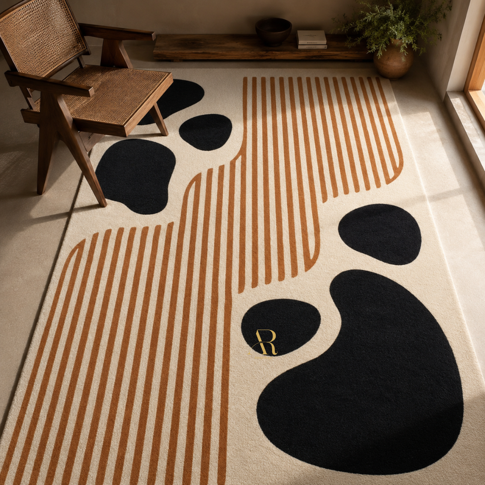 A modern area rug with an abstract design featuring black organic shapes and vertical tan stripes on a cream background, styled in a warm, sunlit room with a wooden chair and shelf.