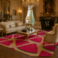 A luxurious living room featuring a large area rug with a bold watermelon pattern in vibrant pink, green, and beige. The room is styled with elegant gold-framed furniture, a crystal chandelier, and a classic fireplace.
