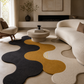 A modern wavy-pattern area rug in charcoal, mustard yellow, and cream, styled in a contemporary living room with a curved cream sofa, a round concrete coffee table, and a minimalist fireplace.