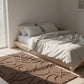 Minimalist bedroom with a wooden platform bed, white bedding, and a textured brown rug.
