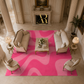 A large rectangular area rug with bold wavy abstract patterns in bright pink and light pink, styled in a luxurious classical living room with beige sofas, armchairs, and side tables.