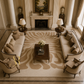 A luxury living room featuring a large beige and brown abstract patterned area rug, coordinated cream sofas and armchairs, dark wood side tables with lamps, and a central coffee table in front of a classical fireplace.