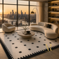 A luxury modern living room featuring a curved cream boucle sofa, a matching armchair, a round stone coffee table, and a large black and cream checkered area rug. The room is set against a backdrop of floor-to-ceiling windows showing a golden city skyline at sunset.