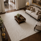 A large cream-colored area rug with a subtle wavy damask pattern, styled in a luxurious, classic living room with a dark wood coffee table, a cream sofa, and dark wood floors.