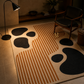 A modern area rug with a beige base, featuring large black organic shapes and rust-orange striped geometric patterns, styled in a warm, dimly lit living room next to a black floor lamp and a wooden armchair.