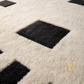 Close-up of a plush rug with a cream background and black geometric diamond pattern, featuring a gold monogram watermark in the lower right corner.