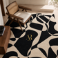 A modern abstract area rug with a bold black and cream wavy pattern, styled in a minimalist interior with a wooden armchair and a small wooden side table. The rug features a gold 'Arredogo' logo in the center.