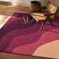 A rectangular area rug with a wavy abstract pattern in shades of magenta, purple, and beige, styled in a sunlit room with a wooden chair and a potted plant.