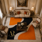 A luxury living room scene featuring a large abstract area rug with orange, black, cream, and white shapes. The rug has a gold 'Artista Rugi' monogram logo in the bottom-left corner.