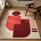 A rectangular area rug with an abstract geometric design in cream, coral red, and deep burgundy, styled on a light wood floor next to a mid-century modern wooden armchair.