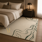 A beige area rug with a dark green wavy line pattern and a gold monogram logo, styled in a modern bedroom next to a bed and a dark wood nightstand.