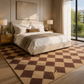 A large checkered area rug in brown and beige tones is styled on the floor of a modern, sunlit bedroom with a bed, bedside tables, lamps, and a large window.